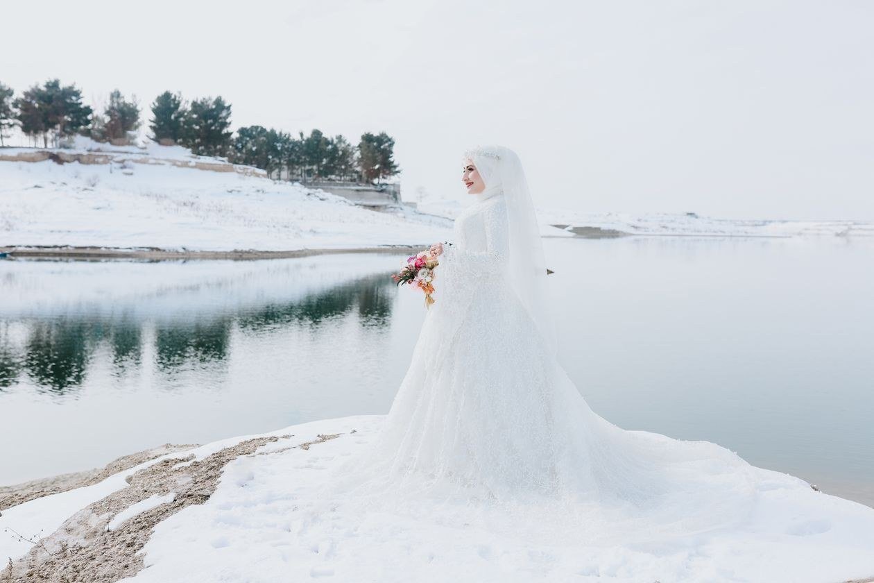 A bride in a white gown stands elegantly by a snow-covered lake, holding a bouquet