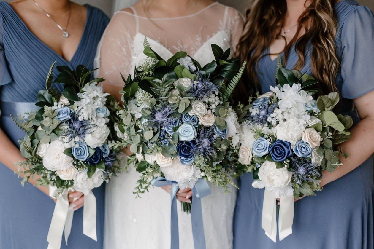 Three women in blue dresses hold elegant white and blue floral bouquets for a wedding